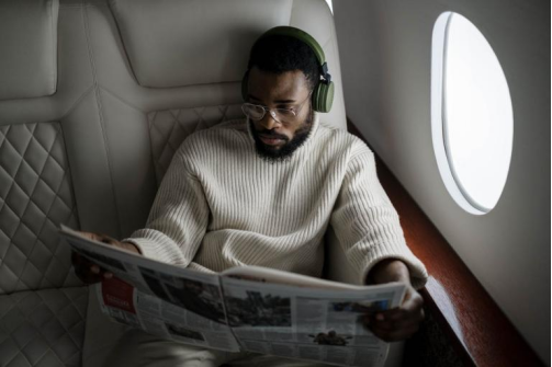 Passenger wearing headphones reading a newspaper while seated in an airplane.