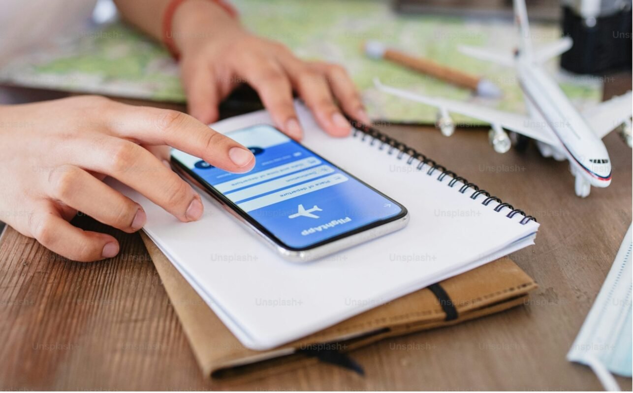 Close-up of a person using a travel booking app on a smartphone, with a notebook and airplane model in the background